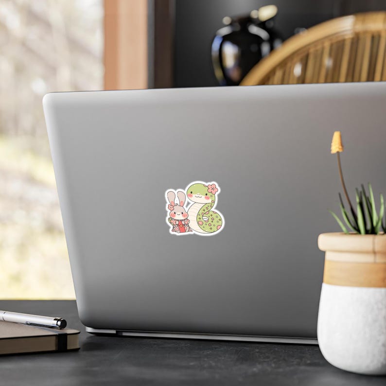 A sticker with a cartoon illustration of a green snake with pink flowers and a white rabbit wearing a pink dress and a pink flower in its hair.