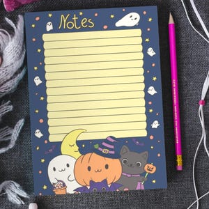 May include: A Halloween notepad with a dark blue background, yellow lined paper, and the word "Notes" at the top. The notepad features illustrations of ghosts, a pumpkin, a black cat, and a crescent moon. A pink pencil is placed on the right.