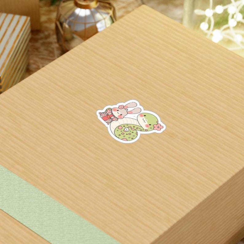 A sticker of a green snake with a pink bunny on its back. The snake and bunny are both cartoonish and have pink flowers on them.