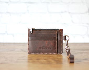 May include: A dark brown leather card holder with a top zipper closure, multiple card slots, and a detachable leather wrist strap. The front features the embossed text THREE TRADESMEN. The wallet has visible light-colored stitching throughout.