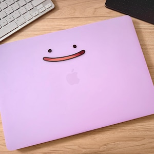 May include: A lilac laptop case with a cartoon face design. The design features two black dots for eyes and a red and pink smiling mouth. The Apple logo is visible. A keyboard and a black tablet are in the background. The case is designed for a laptop.