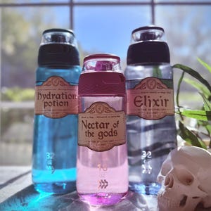 May include: Three water bottles with labels reading "hydration potion", "nectar of the gods", and "elixir". The bottles are filled with blue, pink, and clear liquids. A white skull sits to the right. Each bottle has a capacity of 32 oz or 16 oz.