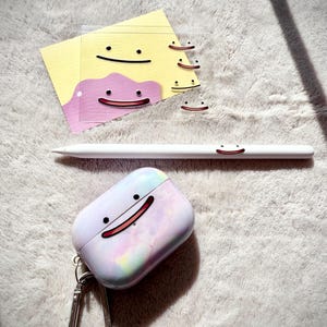 Könnte beinhalten: Ein weißer Stift, ein helllila AirPod-Etui mit einem silbernen Karabiner und ein gelber Haftzettel mit einem rosa und roten Charakterdesign. Das AirPod-Etui hat einen regenbogenfarbenen, schillernden Effekt.
