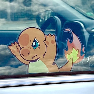 Może przedstawiać: Naklejka z postacią z kreskówki Charmander, z pomarańczową skórą, niebieskimi oczami i płomieniem na ogonie. Naklejka jest przymocowana do szyby samochodu, z wnętrzem widocznym w tle.