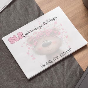 May include: A white notepad featuring the text "SLP Speech-Language Pathologist" and a cartoon bear with a floral crown. The notepad rests on a wooden surface, alongside a laptop, pen, and a potted succulent plant.