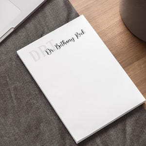 May include: A white notepad with the text "Dr. Bethany Rock" in black script. The notepad is on a dark gray surface next to a laptop and a pen. The notepad is on a wooden surface.