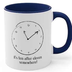 May include: A white ceramic mug with a dark blue interior and handle. The front features a black hand-drawn clock graphic showing the time at ten after eleven, with the text "it's ten after eleven somewhere!" printed in black below the clock face.