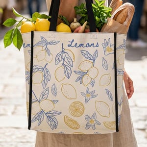 Puede incluir: Un bolso tote de lona crema con asas negras, decorado con un diseño de limones azules y amarillos y la palabra "Lemons". La bolsa está llena de comestibles, incluyendo limones, pan y verduras.