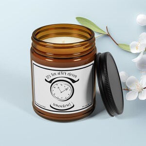 May include: A brown glass jar candle with a black lid, featuring a white label with the text "It's ten after eleven somewhere!" and a clock graphic. The candle is on a light blue surface with white flowers.