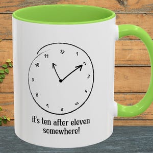 May include: White ceramic mug with a green interior and handle. The mug features a clock design with the text "it's ten after eleven somewhere!" The clock hands point to 11:10.