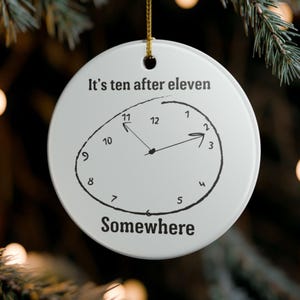 May include: A round white ceramic ornament featuring a hand-drawn black clock face with an irregular circular border. The text "It's ten after eleven" is printed above the clock, and the word "Somewhere" is printed below the clock face.