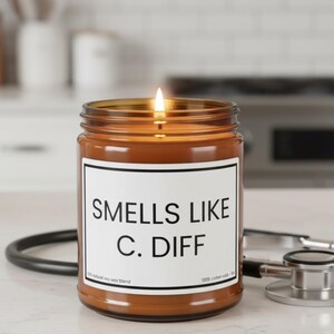 Op de afbeelding: Een brandende bruine glazen potkaars met een wit etiket met de tekst "Smells Like C. Diff." Een zwarte stethoscoop ligt naast de kaars. De kaars staat op een wit oppervlak.