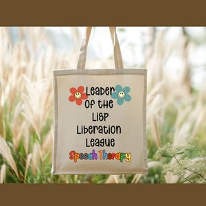 May include: A cream-colored canvas tote bag featuring the black text "Leader of the LiSP Liberation League" with a red flower and a blue flower, each containing a yellow smiley face, and the words "Speech Therapy" in multicolored letters.