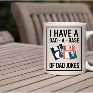 Op de afbeelding: Witte keramische mok met een zwarte rand en tekst die luidt "Ik heb een dad-a-base met papa-grappen". De tekst wordt vergezeld door een afbeelding van drie floppy disks in rood en zwart.