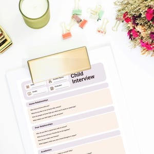 Può includere: Un foglio di carta bianco con un modulo "Child Interview", tenuto da una clip color oro. Il modulo include sezioni per le informazioni sullo studente e domande sui rapporti familiari e con i coetanei, e sugli studi. Sono visibili anche una candela verde, graffette e fiori.
