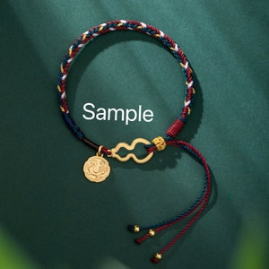 May include: Braided bracelet with red, blue, white, and gold threads. The bracelet features a gold-colored charm with a coin-like pendant and a gourd-shaped connector. The word "Sample" is printed on the image.