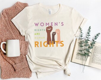 May include: A beige t-shirt with the text "Women's Rights Are Human Rights" in purple, green, and orange. The text is accompanied by a graphic of three raised fists in different skin tones.