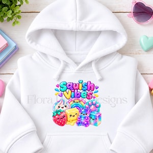 May include: A white hooded sweatshirt featuring a colourful graphic print on the chest. The design includes the text "Squish Vibes" above illustrations of a strawberry, a butter cube, a gummy bear, and other rainbow-coloured shapes.