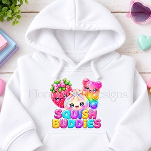May include: A white hooded sweatshirt featuring a colorful graphic of a strawberry, a dumpling, and a gummy bear with smiling faces. Below the characters, the text reads SQUISH BUDDIES in bright, multi-colored bubble letters with a bold outline.