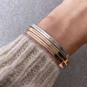 May include: Three gold, silver, and rose gold bracelets stacked on a person's wrist. Each bracelet has the inscription "Everything Happens for my Highest Good" in English and Hebrew.