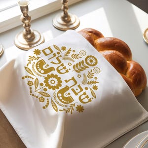 Puede incluir: Una cubierta de challah de tela blanca con un diseño floral dorado y las palabras hebreas "Shabbat Shalom" impresas en ella. La cubierta está drapeada sobre un pan challah trenzado.