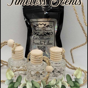 May include: Three glass car air fresheners with wooden tops and a gold string hanging from each. The air fresheners are filled with a clear liquid and have a diamond-shaped pattern. The air fresheners are arranged in a row on a white surface. The image also includes a small bag with the text "Timeless Scents Car diffuser French Lilac" on it.