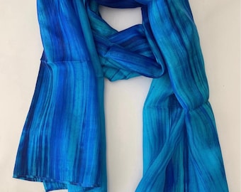 May include: A blue and turquoise silk scarf with a watercolor-like design. The scarf is folded and draped on a white surface.