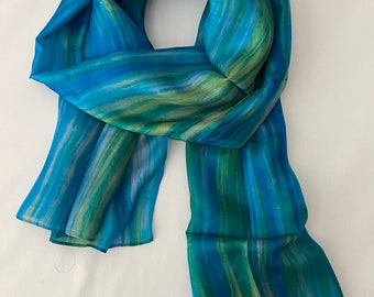 May include: A flowing, rectangular scarf in shades of blue and green. The scarf features a watercolor-like design with vertical stripes of varying widths. The fabric appears to be lightweight and silky, with a subtle sheen. The scarf is draped and folded, showcasing its texture and color variations.