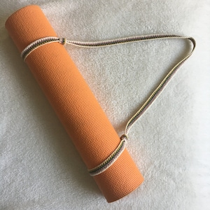 Peut inclure: Un tapis de yoga orange roulé avec une sangle blanche et marron. La sangle est attachée autour du tapis et a un nœud sur le dessus.