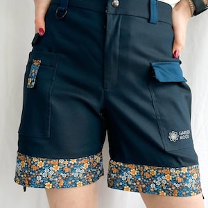 May include: Dark blue utility shorts with a floral print trim. The shorts have two large pockets and a small pocket on the right leg. The right pocket has a white daisy with the text "GARDEN MOOD" embroidered on it.
