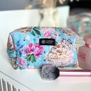 Puede incluir: Una bolsa de maquillaje floral azul claro con una etiqueta negra que dice "Garden Mood".