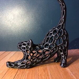 Op de afbeelding: Zwarte, 3D-geprinte kattenfiguur met een traliewerkontwerp. De kat staat in een rekhouding met zijn staart omhoog.