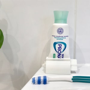 May include: A white toothpaste dispenser with a tube of Prona Sensitive Daily toothpaste inside. The toothpaste tube is blue and white with the text "Prona Sensitive Daily" and "Advanced Enamel Protection" on it. A blue and white toothbrush is on the counter next to the dispenser.