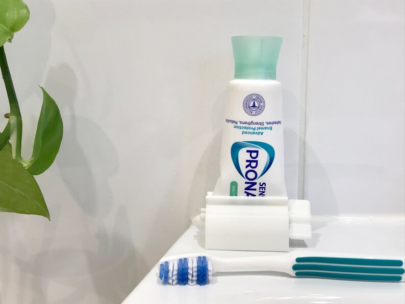 May include: A white toothpaste dispenser with a tube of Prona Sensitive Daily toothpaste inside. The toothpaste tube is blue and white with the text "Prona Sensitive Daily" and "Advanced Enamel Protection" on it. A blue and white toothbrush is on the counter next to the dispenser.