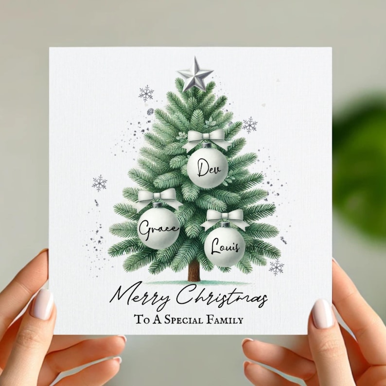 May include: A white Christmas card with a green Christmas tree illustration. The tree has white ornaments with the names "Dev", "Grace", and "Louis" on them. The card reads "Merry Christmas To A Special Family".