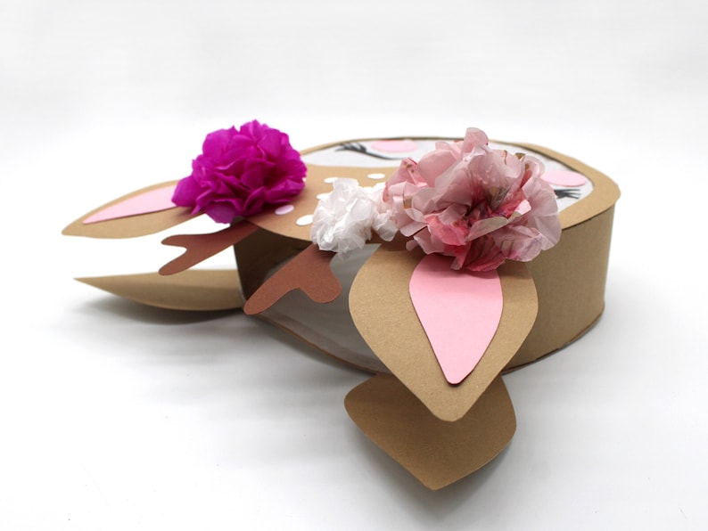 May include: A brown cardboard deer head pinata with pink and white tissue paper flowers. The pinata has a white face with black eyes and eyelashes.