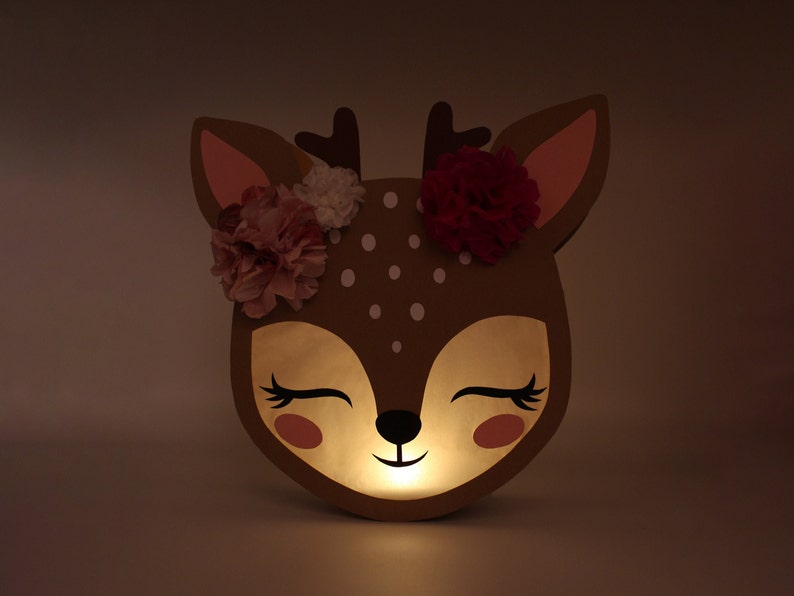 May include: A brown deer-shaped nightlight with a white polka dot pattern and pink and red flowers on its head. The light is on and glowing.
