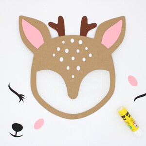 May include: A brown paper deer mask with white polka dots and pink ears. The mask is surrounded by black and pink paper cutouts of eyelashes, a nose, and a heart. A yellow glue stick is also visible in the image.