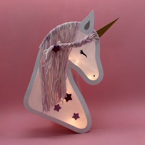 May include: A white unicorn head cutout with a purple and white yarn mane and horn. The unicorn has glitter stars on its face and is illuminated from behind.