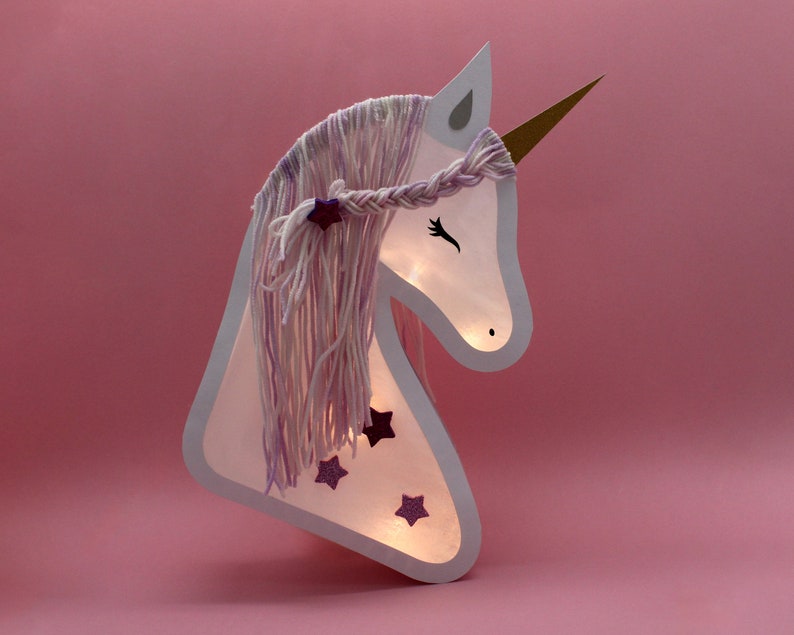 May include: A white unicorn head cutout with a purple and white yarn mane and horn. The unicorn has glitter stars on its face and is illuminated from behind.