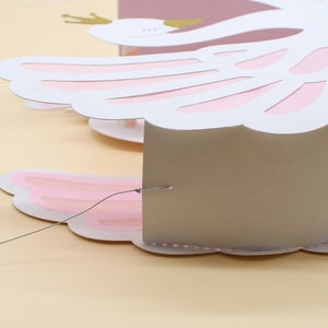 May include: A white paper swan with pink wings and a gold crown. The swan is attached to a thin string and is hanging from the ceiling.