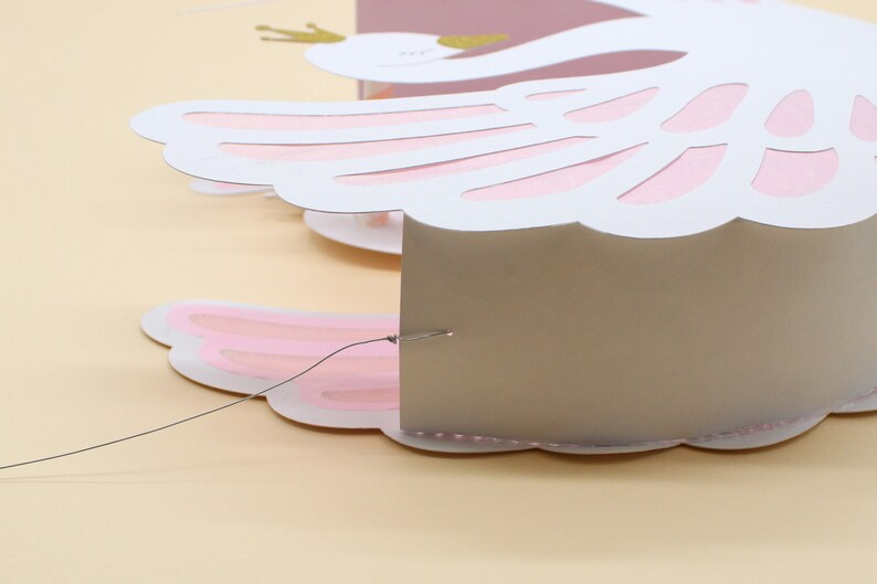 May include: A white paper swan with pink wings and a gold crown. The swan is attached to a thin string and is hanging from the ceiling.