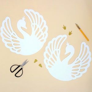 May include: Two white paper swan cutouts with intricate designs, a pair of small scissors, and gold glitter crowns on a light beige background.