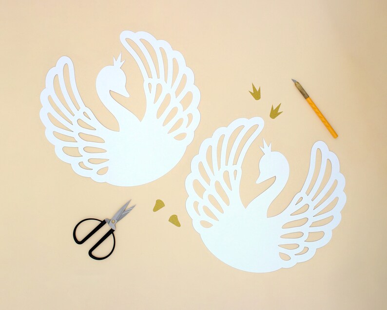 May include: Two white paper swan cutouts with intricate designs, a pair of small scissors, and gold glitter crowns on a light beige background.