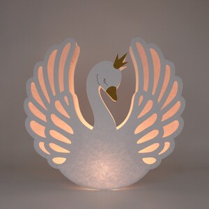 May include: White paper swan lamp with a gold crown and pink wings. The lamp is lit from within, casting a warm glow.