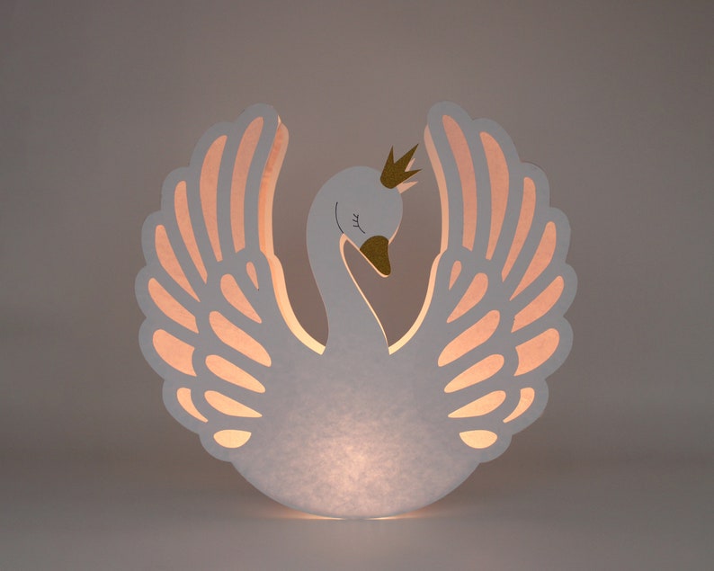 May include: White paper swan lamp with a gold crown and pink wings. The lamp is lit from within, casting a warm glow.