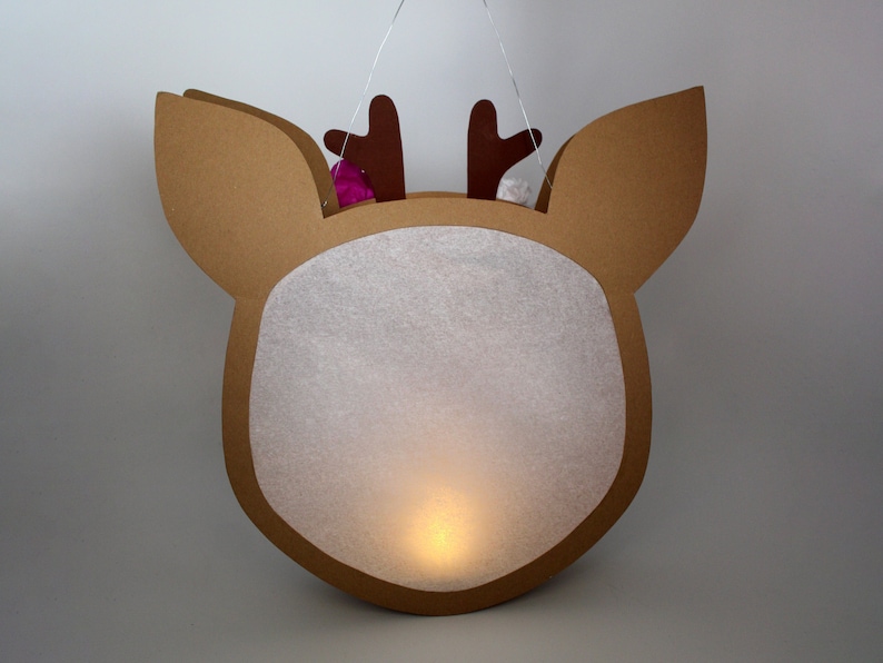 May include: A brown cardboard reindeer-shaped lantern with a white paper insert and a small light inside. The reindeer has brown antlers and a white pom-pom on each antler.