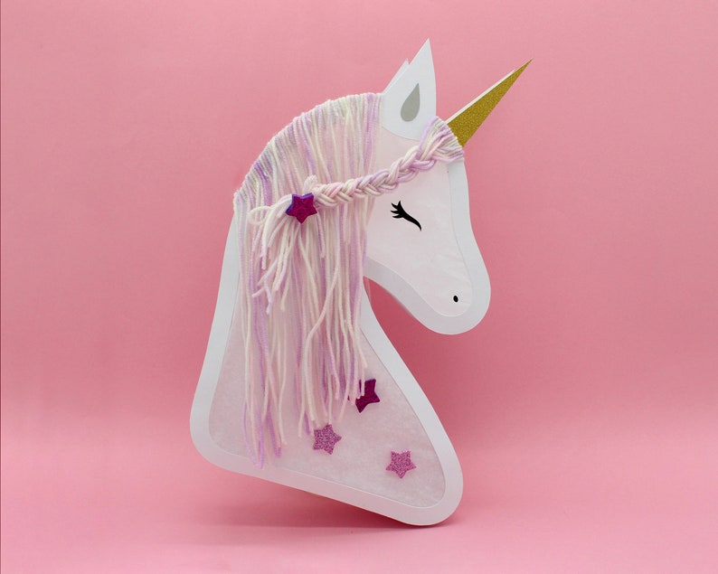 May include: White paper unicorn head with a pink and white yarn mane and horn. The unicorn has pink glitter stars on its face.