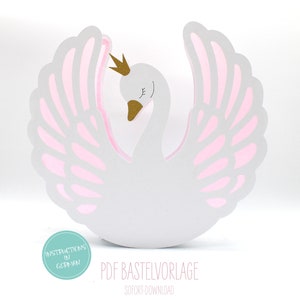 May include: A white papercraft swan with pink wings and a gold crown. The swan is standing on a white surface. The text "PDF Bastevorlage" and "Sofort Download" is printed below the swan.