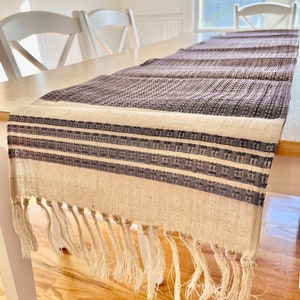 Puede incluir: Un camino de mesa blanco con un patrón tejido gris y blanco y flecos. El camino de mesa está sobre una mesa de madera con dos sillas blancas.